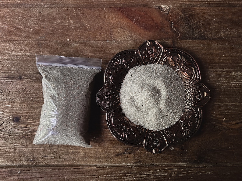 May include: A small plastic bag filled with a gray powder next to a decorative metal dish with a pile of the same gray powder.