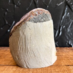 May include: A semi-circular stone specimen featuring a smooth, light gray concrete-like base with visible vertical striations and a crack. The upper portion reveals a natural, rough-textured mineral layer with dark gray and brown crystalline deposits.