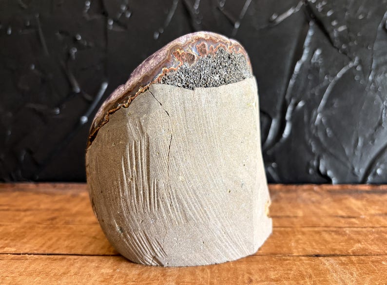 May include: A semi-circular stone specimen featuring a smooth, light gray concrete-like base with visible vertical striations and a crack. The upper portion reveals a natural, rough-textured mineral layer with dark gray and brown crystalline deposits.