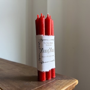 May include: A bundle of red ritual candles wrapped with a white label. The label reads "Ylang Ylang" with the words "Passion & Strength & Courage" below. The candles are on a wooden surface.
