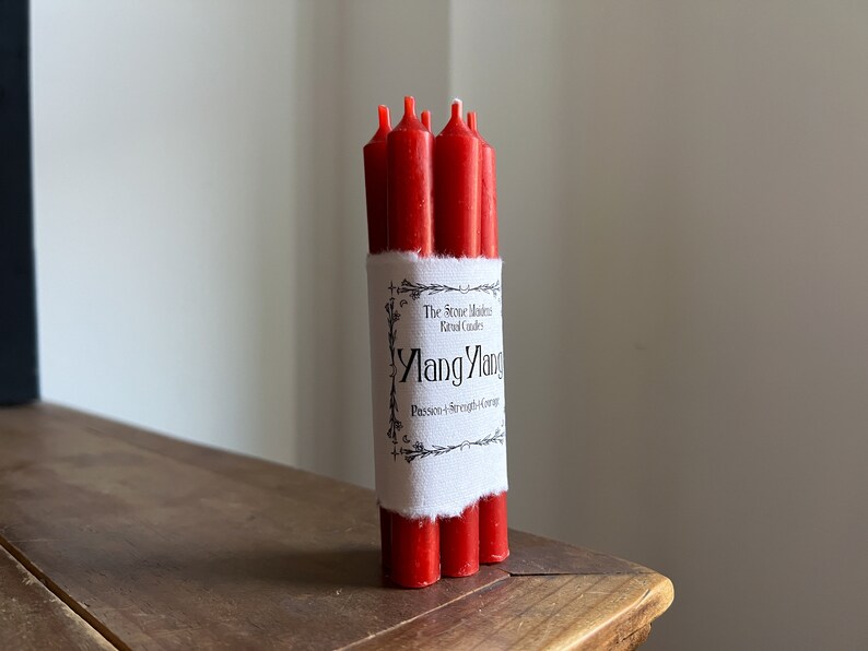 May include: A bundle of red ritual candles wrapped with a white label. The label reads "Ylang Ylang" with the words "Passion & Strength & Courage" below. The candles are on a wooden surface.