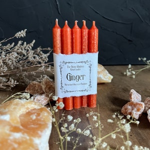K&ouml;nnte beinhalten: Ein Set mit sechs orangefarbenen Ingwer-Ritualkerzen, verpackt in einem wei&szlig;en Etikett mit dem Text "The Stove Maidens Ritual Candles Ginger Attraction + Success + Ambition".