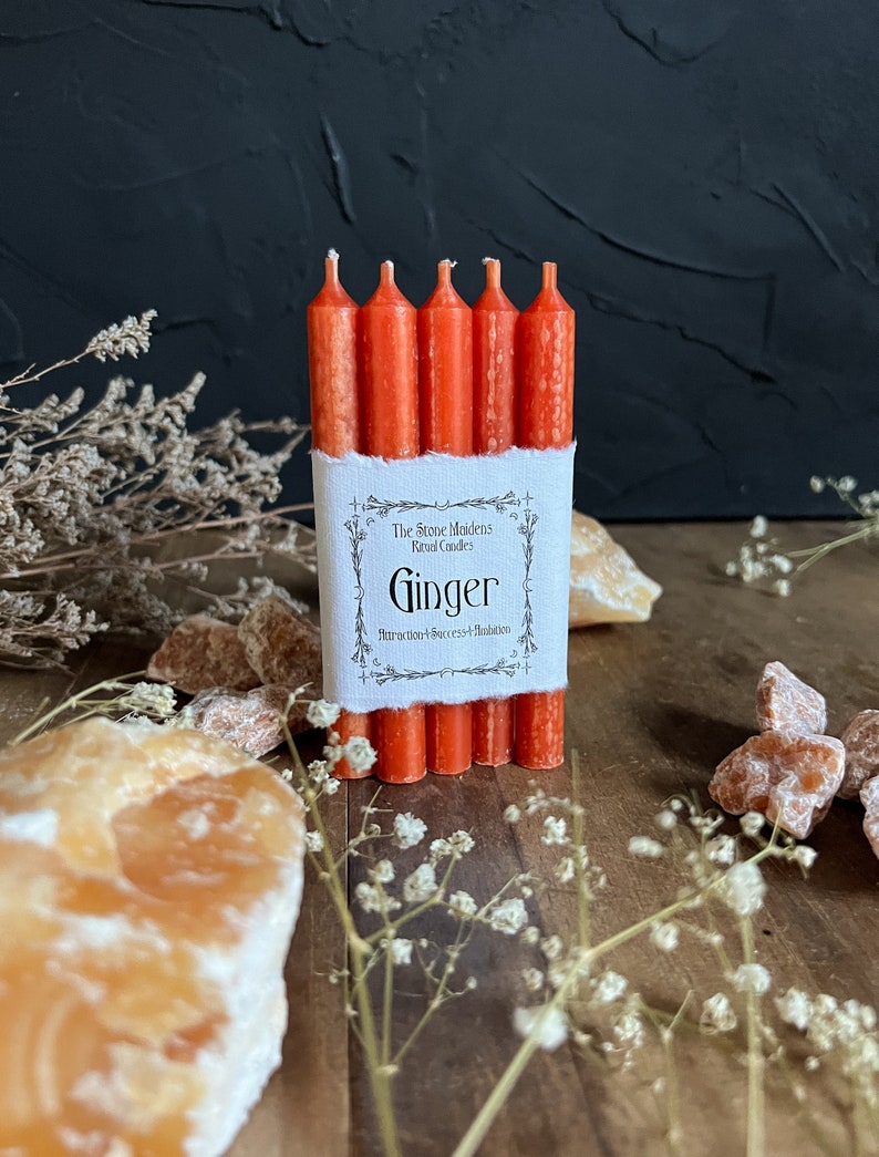 K&ouml;nnte beinhalten: Ein Set mit sechs orangefarbenen Ingwer-Ritualkerzen, verpackt in einem wei&szlig;en Etikett mit dem Text "The Stove Maidens Ritual Candles Ginger Attraction + Success + Ambition".