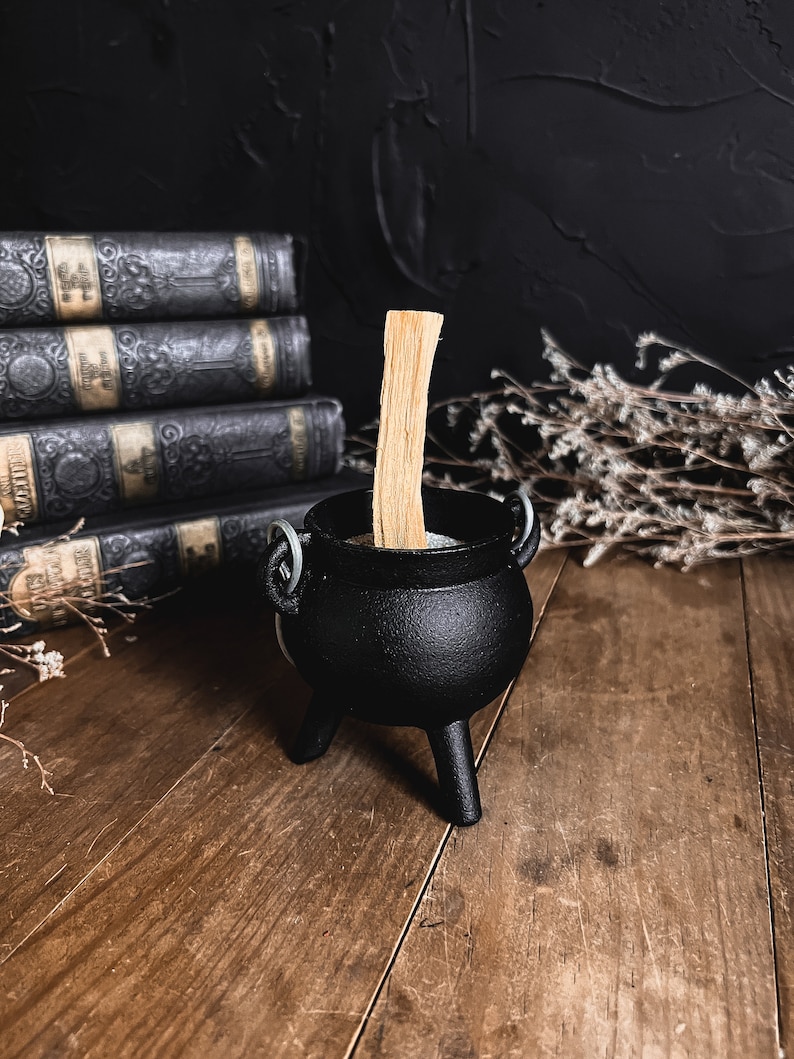 May include: A black cast iron cauldron with three legs and a wooden stick inside. The cauldron is sitting on a wooden surface.