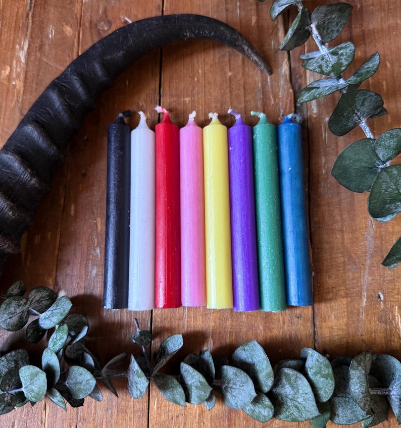 May include: A collection of seven colorful taper candles in a row, with a black candle on the left and a blue candle on the right. A black horn and green leaves are in the background. The candles are various colors, including white, red, pink, yellow, purple, and green.