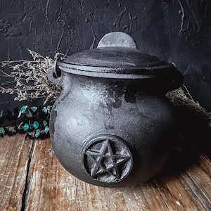 May include: A black cast iron cauldron with a rounded body, a matching lid, and a handle attachment on each side. The front of the cauldron features a raised, embossed pentagram symbol centred on the exterior surface, showcasing a textured finish.