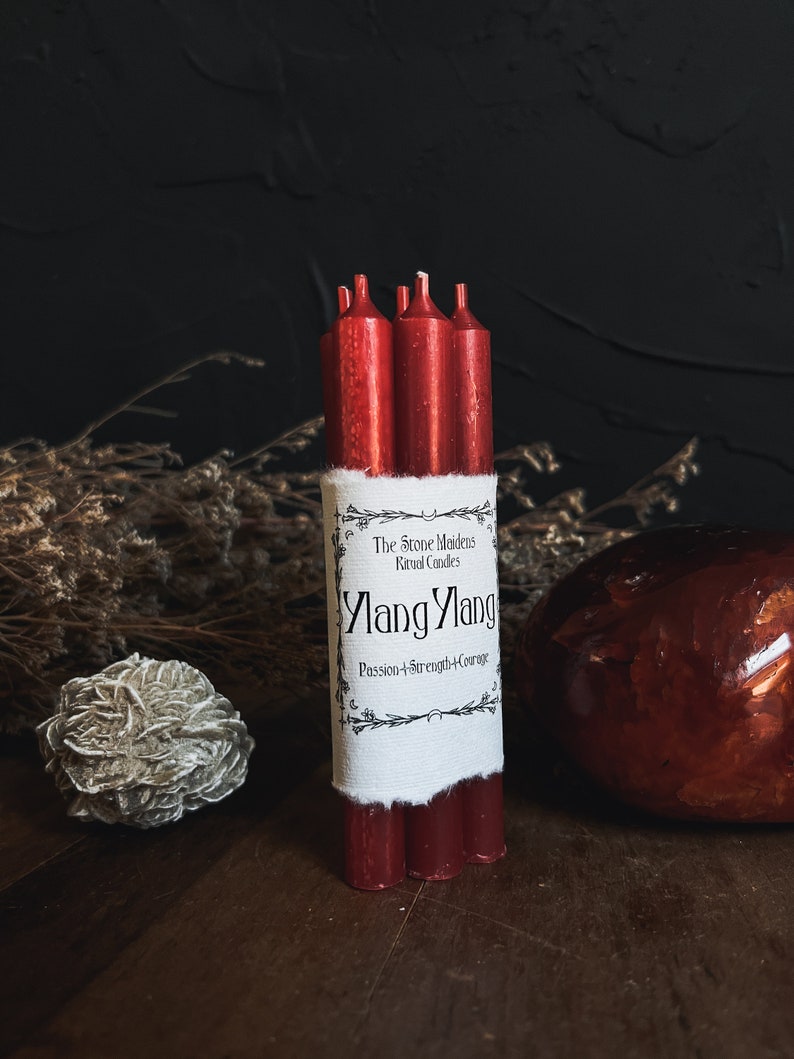 May include: Bundle of red ritual candles with a white label that reads "Ylang Ylang" and "Passion+Strength+Courage". The candles are tied together with a white band. A decorative stone and a dried flower arrangement are in the background.