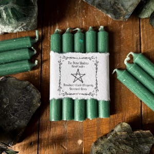 May include: A collection of green ritual candles, with a label reading "The Stone Maidens Ritual Candles" and "Abundance & Luck & Prosperity Unscented Green". The candles are displayed on a wooden surface, alongside decorative stones.