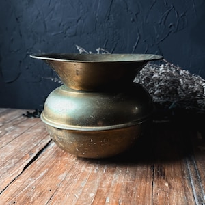 May include: A weathered brass vessel featuring a rounded, bulbous base and a wide, flared rim. The metal surface displays a dark, aged patina with visible signs of oxidation and wear, creating a textured appearance across the entire object.