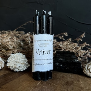 May include: A bundle of three black ritual candles with a white label that reads "Vetiver" and "Protection-Banishing-Grounding". The candles are surrounded by dried plants, crystals, and a black background.