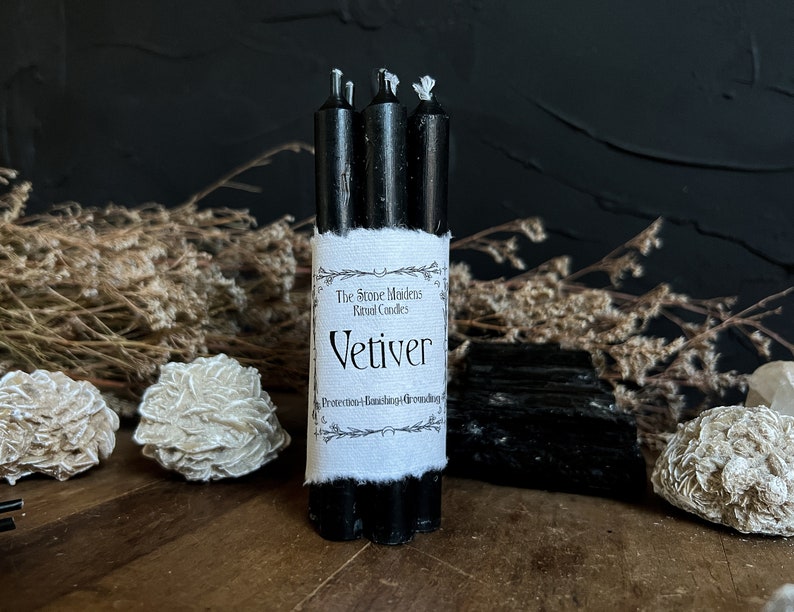May include: A bundle of three black ritual candles with a white label that reads "Vetiver" and "Protection-Banishing-Grounding". The candles are surrounded by dried plants, crystals, and a black background.