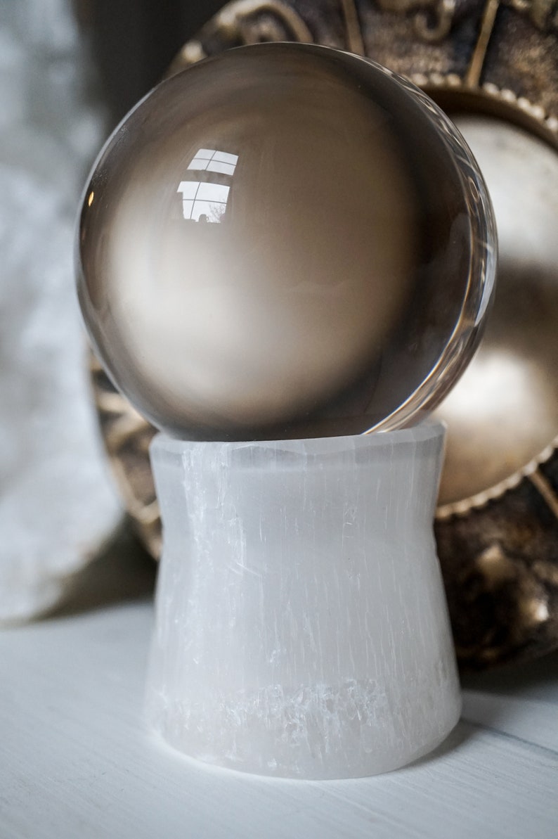 Puede incluir: Una bola de cristal de cuarzo ahumado descansa sobre un pedestal de selenita blanca. La esfera refleja el entorno. El pedestal tiene forma acampanada. El fondo incluye un elemento decorativo.