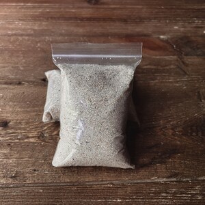May include: A clear plastic bag filled with a light gray sand-like substance. The bag is sealed with a zipper.