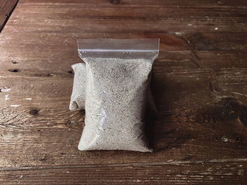 May include: A clear plastic bag filled with a light gray sand-like substance. The bag is sealed with a zipper.