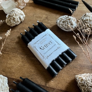 May include: A package of five black vetiver candles. The candles are wrapped in white paper with black text that reads 'The Stone Middens Candles Vetiver Protection, Abundance & Grounding'.