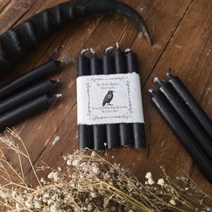 May include: A collection of black ritual candles, some bundled with a white label featuring a raven illustration and text. The candles are arranged on a rustic wooden surface, with dried flowers and a black horn in the background.