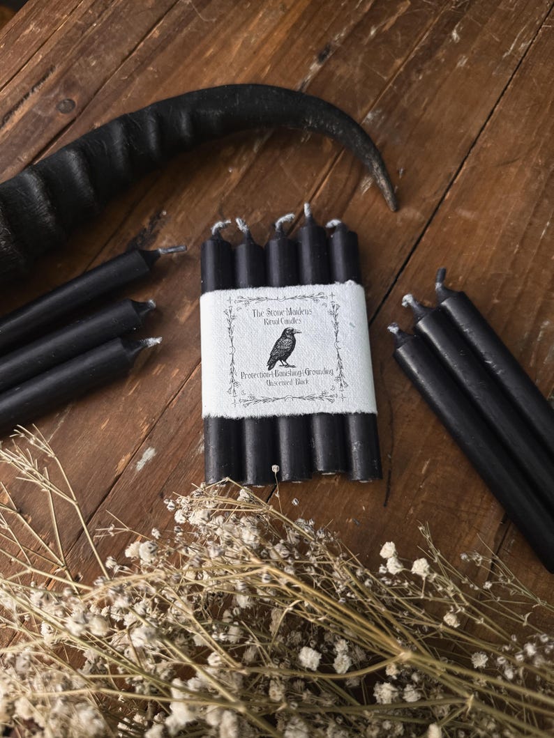 May include: A collection of black ritual candles, some bundled with a white label featuring a raven illustration and text. The candles are arranged on a rustic wooden surface, with dried flowers and a black horn in the background.
