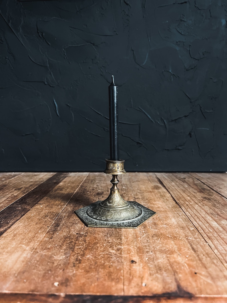 May include: A vintage brass candlestick with a black candle. The hexagonal base and stem feature intricate floral patterns. The candleholder is set on a wooden surface against a dark textured background.