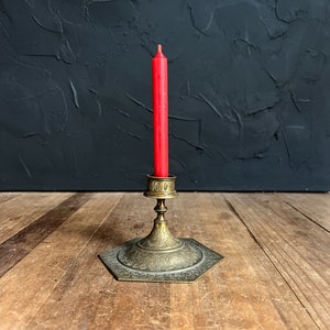 May include: A red taper candle in a vintage brass candlestick holder. The hexagonal base and stem of the holder are adorned with etched floral patterns. The candleholder is on a wooden surface with a dark textured background.