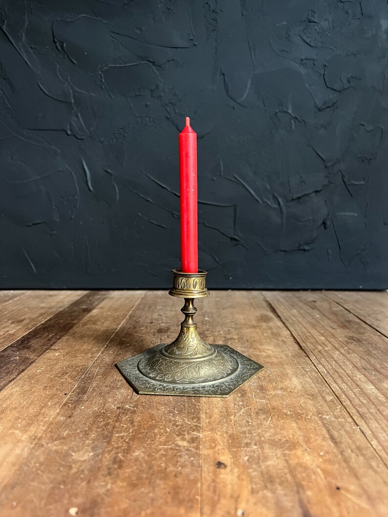 May include: A red taper candle in a vintage brass candlestick holder. The hexagonal base and stem of the holder are adorned with etched floral patterns. The candleholder is on a wooden surface with a dark textured background.
