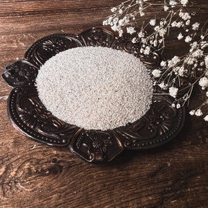 May include: A white, round, decorative plate with a raised, ornate design sits on a brown wooden surface. The plate is filled with white sand or small white beads. A sprig of white flowers is placed on the right side of the plate.