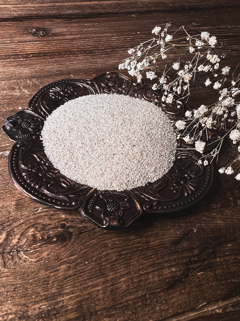 May include: A white, round, decorative plate with a raised, ornate design sits on a brown wooden surface. The plate is filled with white sand or small white beads. A sprig of white flowers is placed on the right side of the plate.