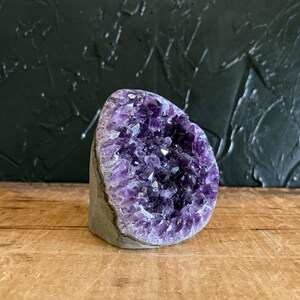 May include: A raw amethyst geode featuring a cluster of deep purple, multifaceted crystals set within a natural grey stone exterior. The specimen is shaped like a rounded wedge, showcasing a dense formation of jagged, translucent violet points.