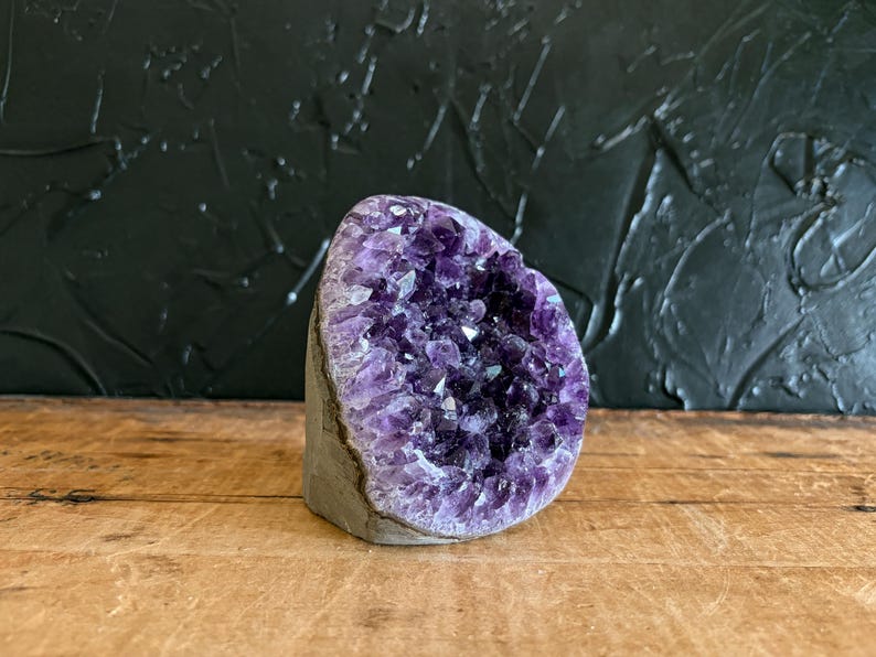 May include: A raw amethyst geode featuring a cluster of deep purple, multifaceted crystals set within a natural grey stone exterior. The specimen is shaped like a rounded wedge, showcasing a dense formation of jagged, translucent violet points.