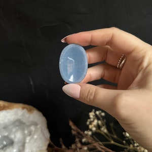 May include: A smooth, oval-shaped blue celestite crystal palm stone. The stone is translucent and has a light blue color.