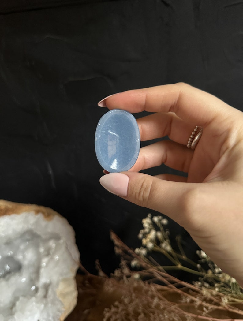 May include: A smooth, oval-shaped blue celestite crystal palm stone. The stone is translucent and has a light blue color.