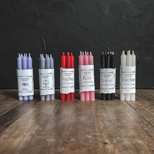 May include: Bundles of ritual candles in various colors, including lavender, gray, red, pink, black, and white. Each bundle is wrapped with a label that includes text. The candles are arranged on a wooden surface against a dark background.