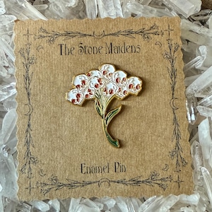 Może przedstawiać: Emaliowana przypinka w kolorze złotym przedstawiająca bukiet białych czaszek z czerwonymi akcentami i zielonymi łodygami. Przypinka umieszczona jest na brązowej karcie z napisem "The Stone Maidens" i "Enamel Pin".