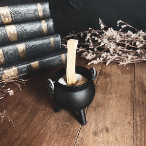 May include: A black cast iron cauldron with three legs sits on a wooden surface. The cauldron is filled with white sand and a wooden stick is placed upright in the sand.