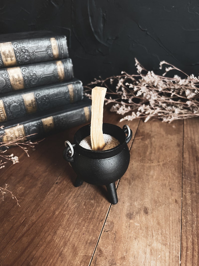 May include: A black cast iron cauldron with three legs sits on a wooden surface. The cauldron is filled with white sand and a wooden stick is placed upright in the sand.