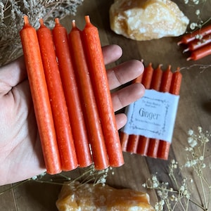 K&ouml;nnte beinhalten: Eine Packung mit sechs orangefarbenen, nach Ingwer duftenden Kerzen. Die Kerzen sind etwa 10 cm lang und in einer wei&szlig;en Papierverpackung mit dem Aufdruck "The Scent Makers Ginger" verpackt.