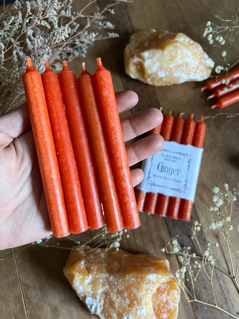 K&ouml;nnte beinhalten: Eine Packung mit sechs orangefarbenen, nach Ingwer duftenden Kerzen. Die Kerzen sind etwa 10 cm lang und in einer wei&szlig;en Papierverpackung mit dem Aufdruck "The Scent Makers Ginger" verpackt.