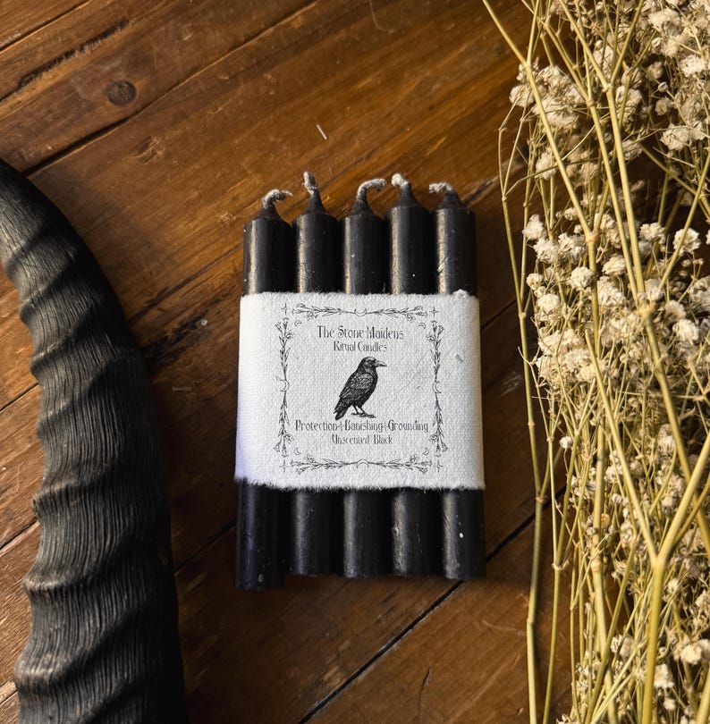 May include: A bundle of black ritual candles with a white label featuring a raven illustration and text. The candles are unscented and designed for protection, banishing, and grounding. The candles are on a wooden surface.