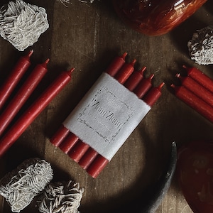 May include: Red taper candles, some bundled with a paper label that reads "Ylang Ylang". The candles are arranged on a wooden surface with decorative stones and crystals. The overall aesthetic is rustic and natural.