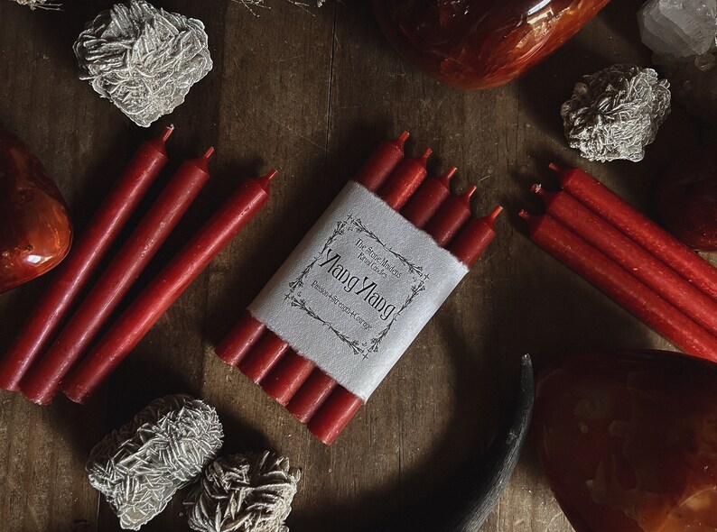 May include: Red taper candles, some bundled with a paper label that reads "Ylang Ylang". The candles are arranged on a wooden surface with decorative stones and crystals. The overall aesthetic is rustic and natural.
