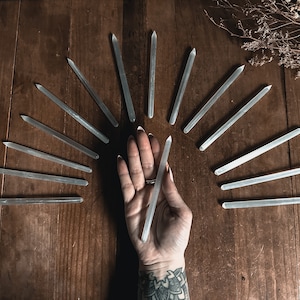 May include: A collection of clear quartz crystal wands arranged in a semi-circle on a wooden surface. A hand is holding one of the wands in the centre of the arrangement.