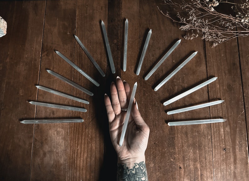 May include: A collection of clear quartz crystal wands arranged in a semi-circle on a wooden surface. A hand is holding one of the wands in the centre of the arrangement.