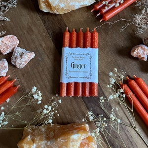K&ouml;nnte beinhalten: Ein Set mit sechs roten Ingwerkerzen mit einem wei&szlig;en Etikett, auf dem "The Stone Maidens Ritual Candles Ginger Attracts Success & Abundance" steht.