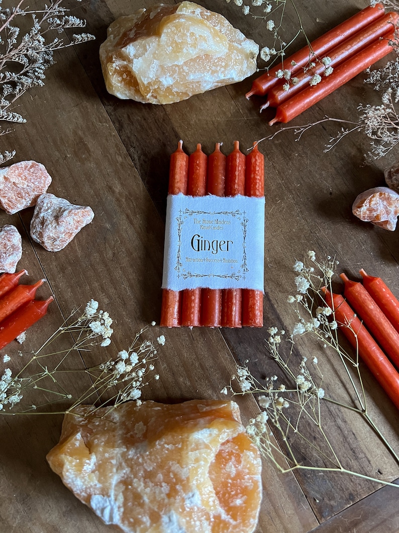 K&ouml;nnte beinhalten: Ein Set mit sechs roten Ingwerkerzen mit einem wei&szlig;en Etikett, auf dem "The Stone Maidens Ritual Candles Ginger Attracts Success & Abundance" steht.