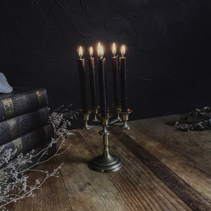 May include: A vintage brass candelabra with six lit black candles. The candelabra sits on a wooden surface, with a stack of antique books and dried flowers in the background. The flames cast a warm glow against the dark backdrop.