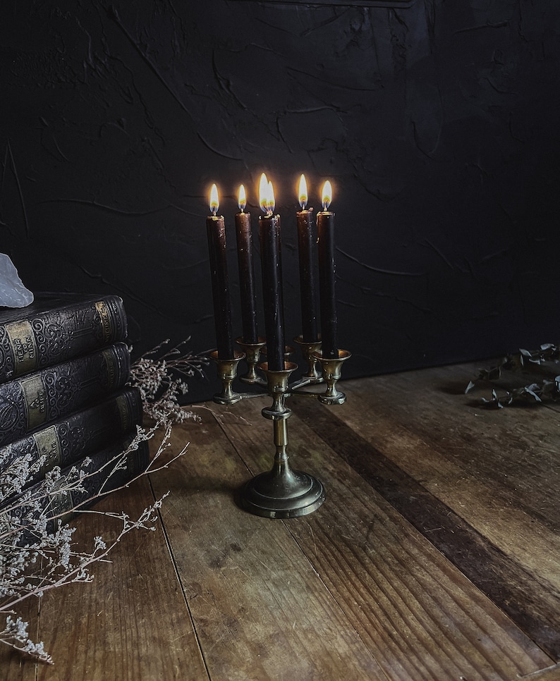 May include: A vintage brass candelabra with six lit black candles. The candelabra sits on a wooden surface, with a stack of antique books and dried flowers in the background. The flames cast a warm glow against the dark backdrop.