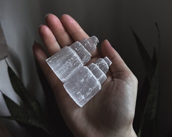 May include: Two white selenite crystal clusters, also known as desert rose crystals, are held in a hand. The crystals are stacked on top of each other and have a layered, striated appearance.