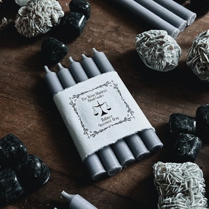 May include: Six grey unscented candles bundled together with a white label that reads "The Stone Magick Hand Candles Balance Unscented Grey". The label features a black scale design and a floral border.