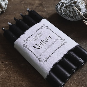 May include: A bundle of black ritual candles wrapped in a white paper label. The label reads "Vetiver" with text about protection, banishing, and grounding. The candles are on a wooden surface.