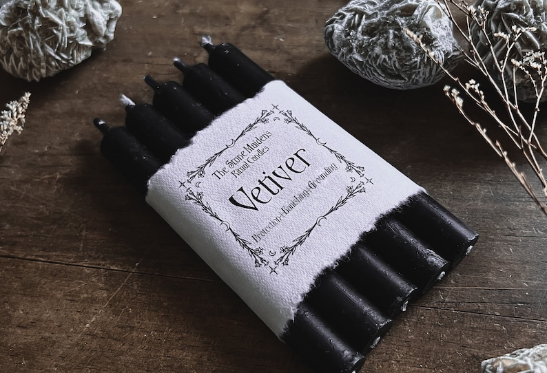 May include: A bundle of black ritual candles wrapped in a white paper label. The label reads "Vetiver" with text about protection, banishing, and grounding. The candles are on a wooden surface.