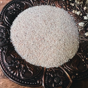 May include: A white, fine-grained powder sits in a decorative, ornate, bronze-colored dish with floral designs. The dish is on a wooden surface.